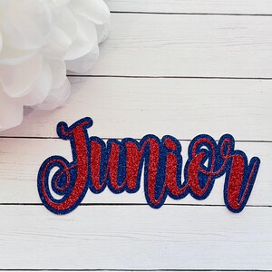 May include: Red and blue glitter letters spelling out the word "Junior" on a white wooden background.