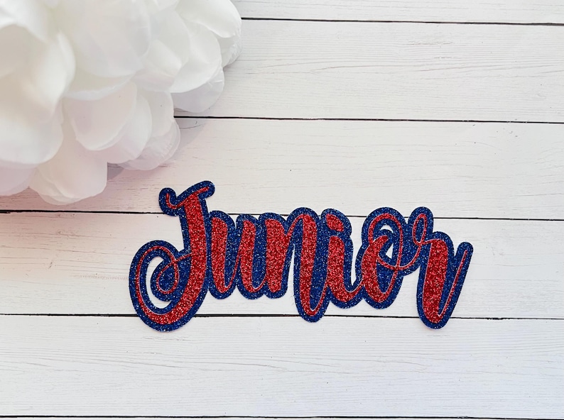 May include: Red and blue glitter letters spelling out the word "Junior" on a white wooden background.