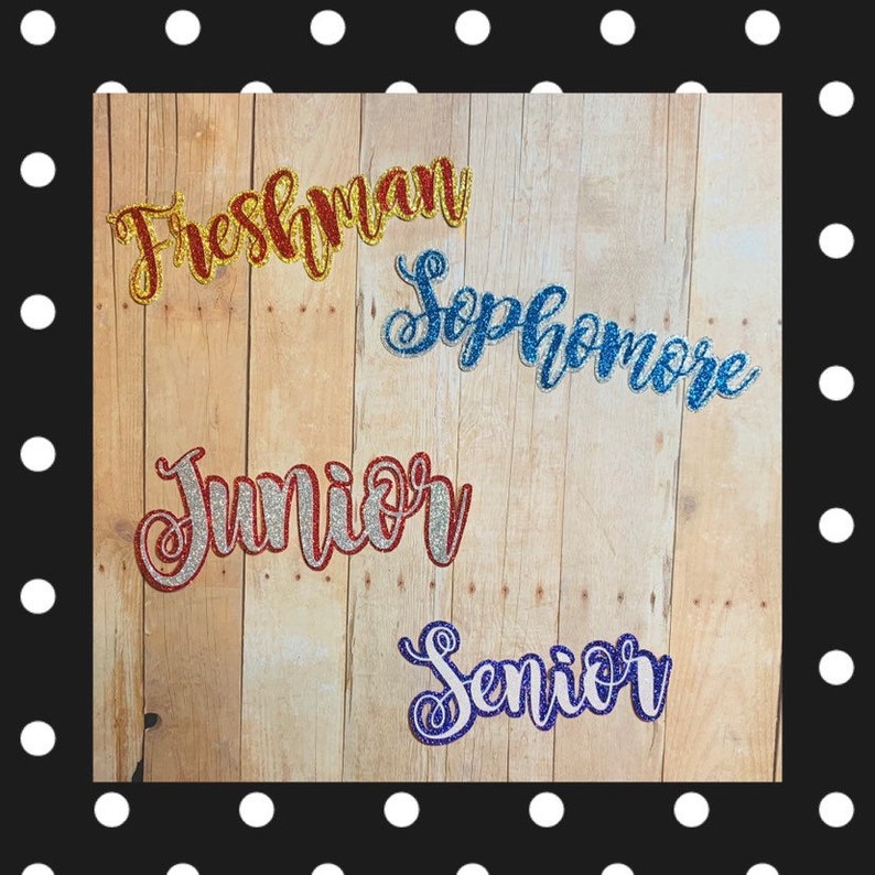 May include: Four glitter iron-on patches with the words "Freshman", "Sophomore", "Junior", and "Senior" in different colors. The patches are on a wooden background.