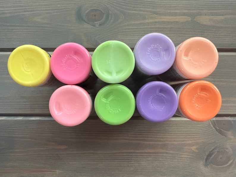 Peut inclure: Huit petits flacons de peinture de couleurs pastel diff&eacute;rentes, dont jaune, rose, vert, violet et orange. Les flacons sont dispos&eacute;s en rang&eacute;e sur une surface en bois.