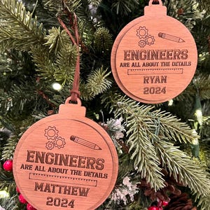 May include: Two wooden Christmas ornaments with a brown wood grain finish. The ornaments are round and feature a pencil, gears, and a ruler. The text on the ornaments reads "Engineers are all about the details". One ornament has the name "Matthew" and the year "2024" on it. The other ornament has the name "Ryan" and the year "2024" on it.
