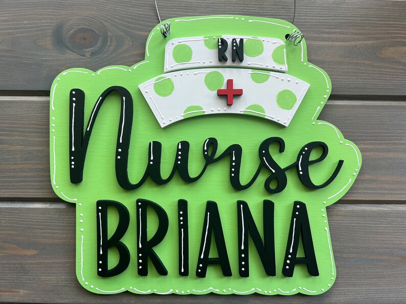 Peut inclure: Une pancarte en bois verte avec des pois blancs et le texte "Nurse Briana" en noir. Un chapeau d'infirmi&egrave;re blanc avec une croix rouge et les lettres "RN" est sur le dessus de la pancarte.