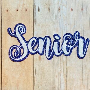 May include: A white glitter "Senior" sign with a blue outline. The sign is made of thick cardstock and is perfect for a graduation party or celebration.