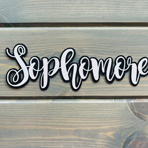 May include: White glitter "Sophomore" lettering on a black background. The lettering is cut out and has a slightly raised texture.