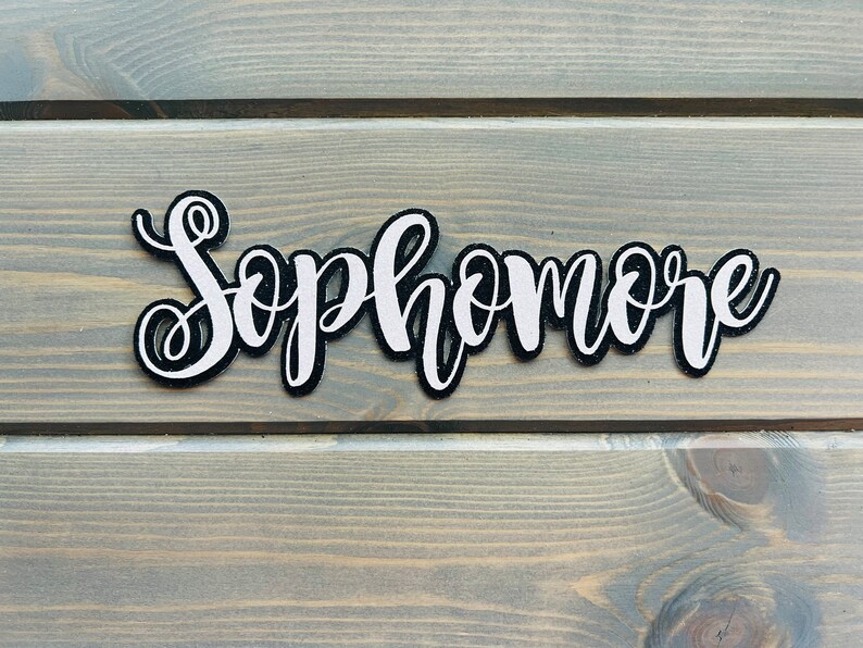 May include: White glitter "Sophomore" lettering on a black background. The lettering is cut out and has a slightly raised texture.