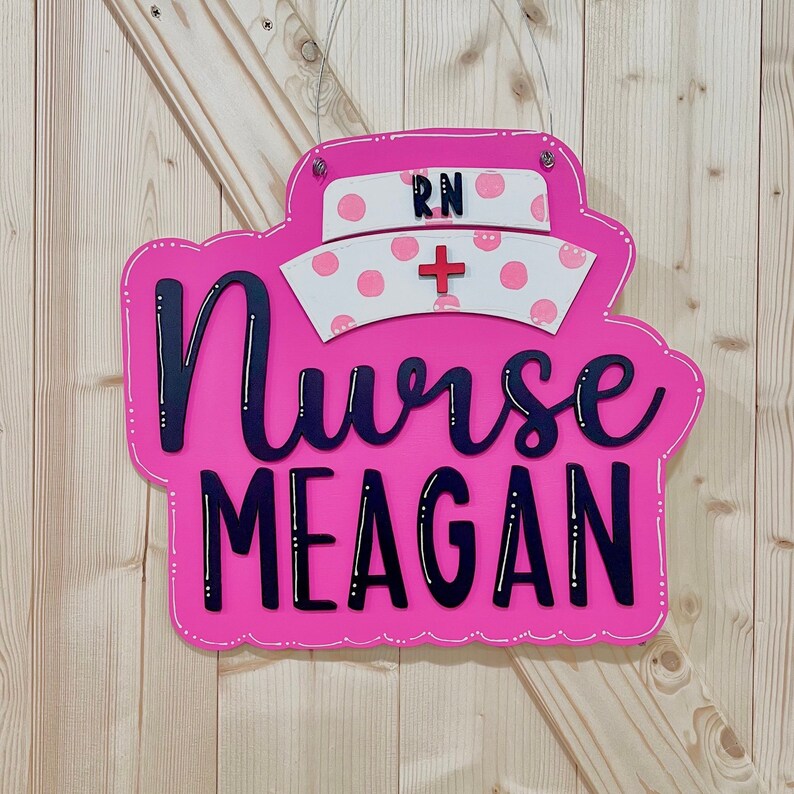 Peut inclure: Une pancarte en bois rose avec un chapeau d'infirmi&egrave;re blanc et rose avec le texte "RN" et une croix rouge. La pancarte indique "Nurse Meagan" en lettres noires.