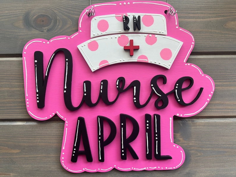 Peut inclure: Une pancarte en bois rose avec des pois blancs et le texte "Nurse April" en lettres noires. Un chapeau d'infirmi&egrave;re blanc avec des pois roses et les lettres "RN" en noir est plac&eacute; sur le dessus de la pancarte.