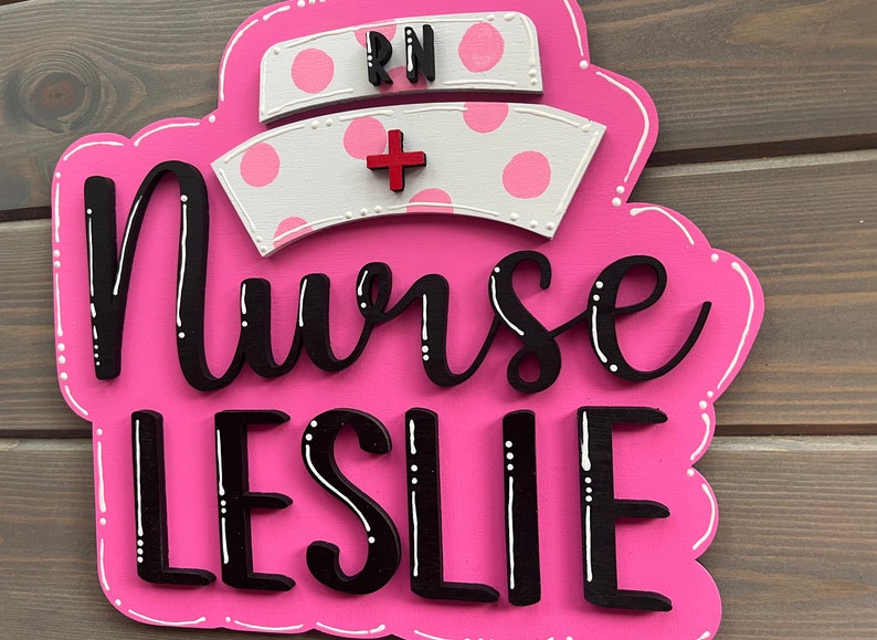 Peut inclure: Une pancarte en bois rose avec des pois blancs et le texte "Nurse LESLIE" en lettres noires. Un chapeau d'infirmi&egrave;re blanc avec des pois roses et les lettres "RN" en noir est au-dessus du texte.