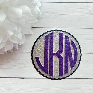 May include: A circular scalloped-edge patch featuring a silver glitter background with a large, bold, purple glitter monogram of the letters JKN centered on the surface. The design is layered with a dark border outlining the scalloped shape.