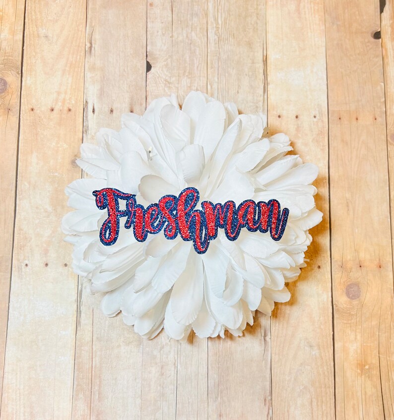 May include: A white paper flower with the word "Freshman" in red and blue glitter letters.