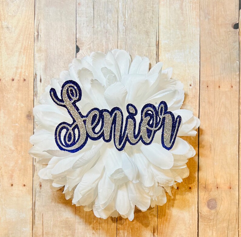 May include: A white paper flower with a silver glitter and navy blue outline "Senior" sign.
