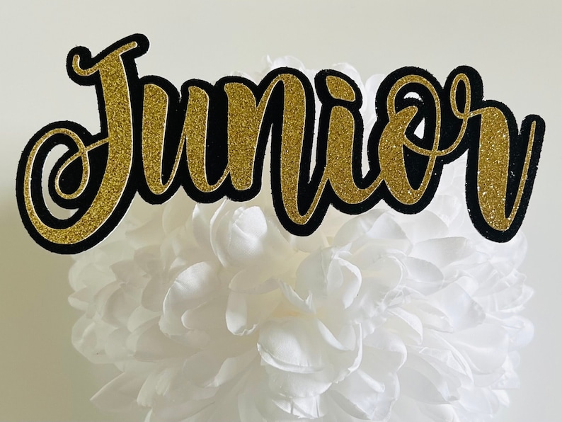 May include: Black and gold glitter cake topper with the word "Junior" in a cursive font.