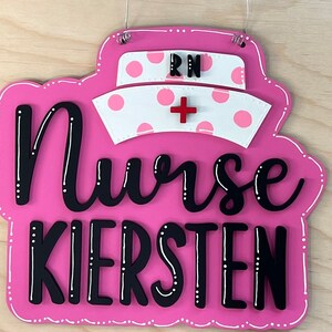Peut inclure: Une pancarte en bois rose avec des points blancs et une croix rouge. La pancarte indique "Nurse Kiersten" et "RN" en lettres noires.