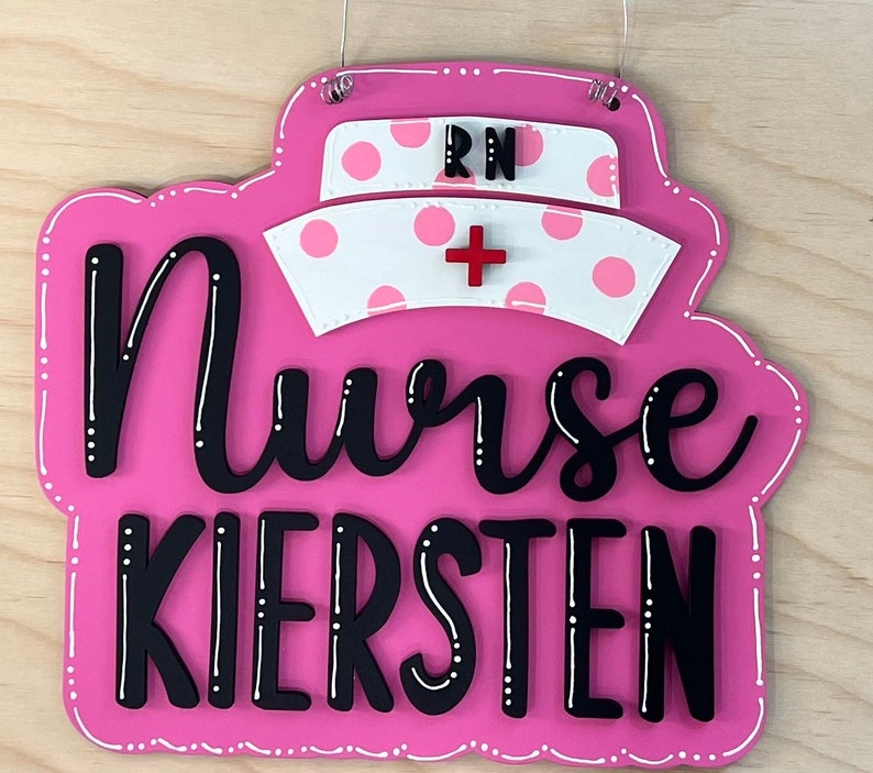 Peut inclure: Une pancarte en bois rose avec des points blancs et une croix rouge. La pancarte indique "Nurse Kiersten" et "RN" en lettres noires.