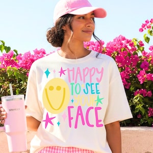 May include: A cream-colored t-shirt featuring a yellow smiley face and the text "HAPPY TO SEE YOUR FACE" in pink and teal, accented with star shapes. The shirt is paired with a pink baseball cap and a light pink insulated tumbler with a straw.