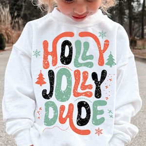 May include: A white long-sleeve sweatshirt featuring a graphic print with the text "HOLLY JOLLY DUDE" in a distressed, multi-colored font of orange, black, and sage green, accented with small illustrations of snowflakes and Christmas trees.