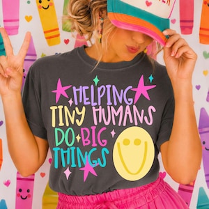 Może przedstawiać: Ciemnoszary t-shirt z kolorowym napisem "HELPING TINY HUMANS DO BIG THINGS", żółtą buźką i różowymi gwiazdkami. Na głowie znajduje się kremowo-turkusowa czapka z daszkiem z napisem "TEACHER" wykonanym tęczowymi literami.