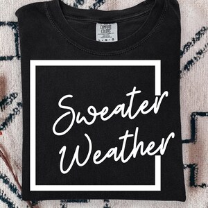 May include: A black crew neck t-shirt featuring a white square outline graphic with the text Sweater Weather written in a white cursive script font across the centre. The shirt has a Comfort Colors brand label at the inner neckline showing size L.