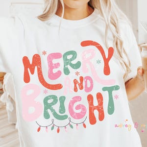 以下が含まれることがあります： 白いTシャツに、赤、緑、ピンク、白のグラフィックで「メリー＆ブライト」と書かれており、その下にクリスマスの電飾が描かれています。