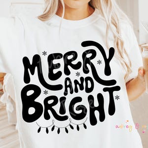 以下が含まれることがあります： 白いTシャツに黒い文字で「メリーアンドブライト」と書いてあり、その下に電飾の紐があります。文字は楽しくてお祝いの雰囲気のフォントです。