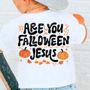 May include: A white t-shirt featuring black text that reads Are You Falloween Jesus. The design includes orange pumpkins, candy corn, and small star and lightning bolt icons. The shirt is worn with blue denim jeans and a gray baseball cap.
