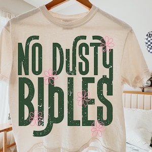 May include: A cream-colored t-shirt with the text "No Dusty Bibles" in dark green, accented with pink flower graphics. The shirt is hanging on a wooden hanger.