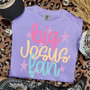 K&ouml;nnte beinhalten: Ein lila Comfort Colors T-Shirt mit den Worten "big Jesus fan" in Pink, Gelb und T&uuml;rkis, mit rosa Sternakzenten. Das Shirt wird auf einer Oberfl&auml;che mit Leopardenmuster pr&auml;sentiert.