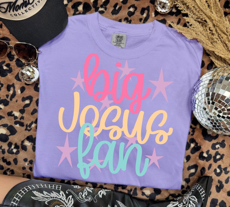 K&ouml;nnte beinhalten: Ein lila Comfort Colors T-Shirt mit den Worten "big Jesus fan" in Pink, Gelb und T&uuml;rkis, mit rosa Sternakzenten. Das Shirt wird auf einer Oberfl&auml;che mit Leopardenmuster pr&auml;sentiert.