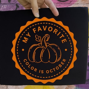 May include: A black t-shirt featuring a circular orange graphic with a scalloped edge. The design includes a central pumpkin illustration surrounded by the text MY FAVORITE COLOR IS OCTOBER in a bold, sans-serif font within the circular border.