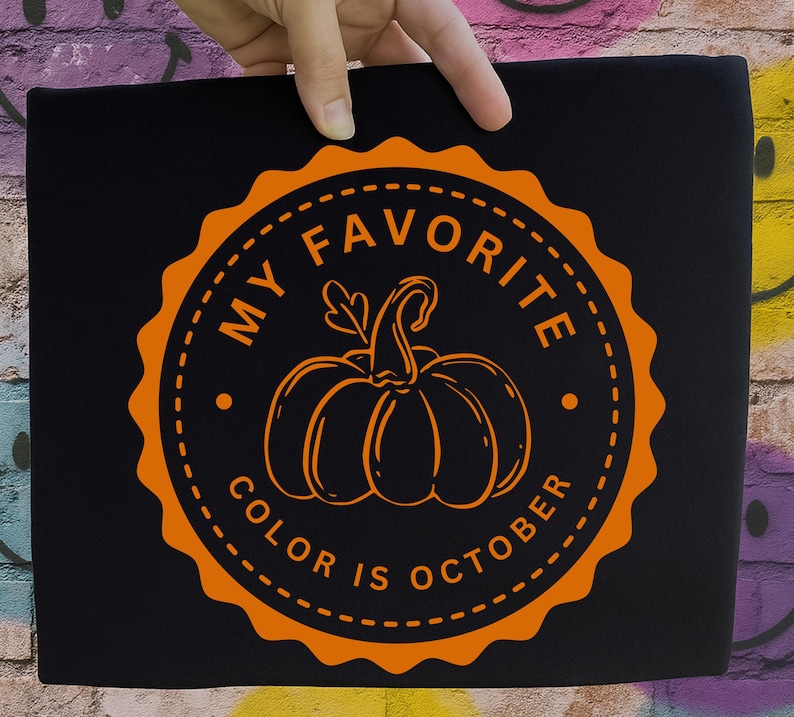 May include: A black t-shirt featuring a circular orange graphic with a scalloped edge. The design includes a central pumpkin illustration surrounded by the text MY FAVORITE COLOR IS OCTOBER in a bold, sans-serif font within the circular border.