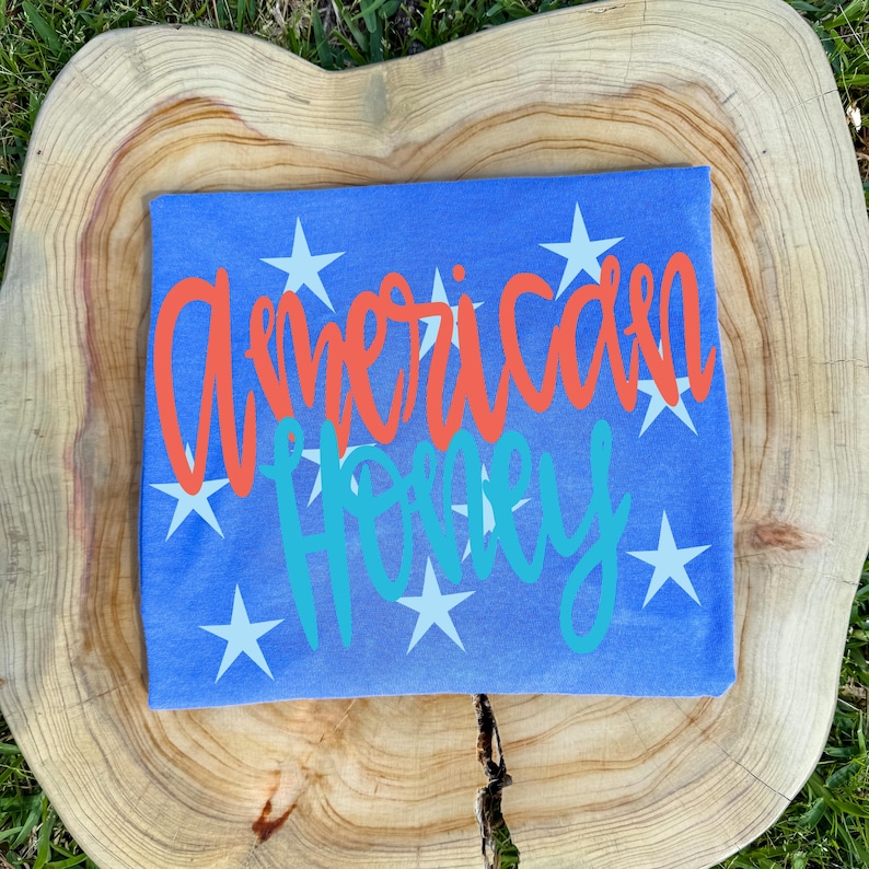 May include: A blue t-shirt with the words "American Honey" in orange and teal script, surrounded by white stars. The shirt is displayed on a wooden surface.