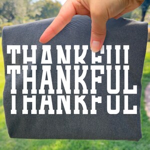 May include: A dark charcoal grey t-shirt featuring the word THANKFUL printed in large, bold, white block letters across the chest. The text is repeated in three horizontal rows, with the word THANKFUL appearing in a clean, sans-serif typeface.