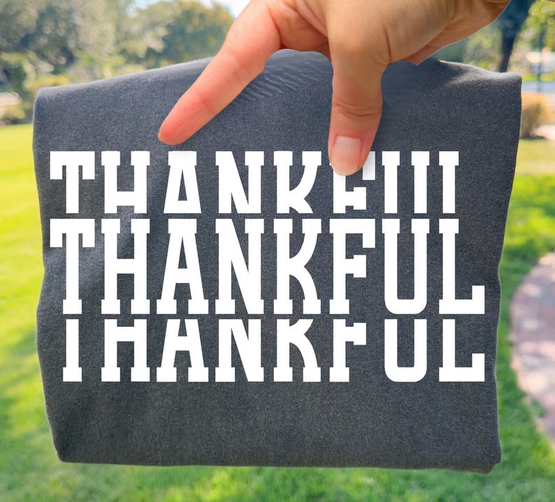May include: A dark charcoal grey t-shirt featuring the word THANKFUL printed in large, bold, white block letters across the chest. The text is repeated in three horizontal rows, with the word THANKFUL appearing in a clean, sans-serif typeface.