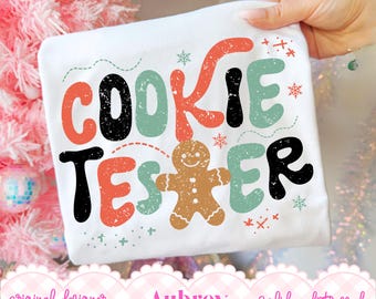 以下が含まれることがあります： 赤、黒、セージグリーンの掠れたマルチカラーフォントで「COOKIE TESTER」とプリントされた白いTシャツ。TESTERの「I」の文字が茶色のジンジャーブレッドマンのアイコンに置き換わっており、周囲には小さな雪の結晶のアクセントが散りばめられています。