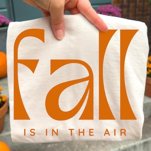 May include: A white t-shirt featuring the word fall in large, bold, orange stylised lettering. Below the word fall, the text IS IN THE AIR is printed in a smaller, simple orange font. The design is centred on the front of the garment.