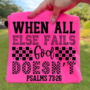 May include: A vibrant pink t-shirt featuring the phrase "WHEN ALL ELSE FAILS GOD DOESN'T" in black. The words "ELSE FAILS" have a leopard print design. The text "PSALMS 73:26" is printed at the bottom of the shirt.