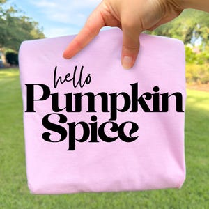May include: A light pink t-shirt featuring the black text "hello Pumpkin Spice" printed on the front. The word "hello" is written in a cursive script font, while "Pumpkin Spice" is displayed in a bold, stylized serif typeface.