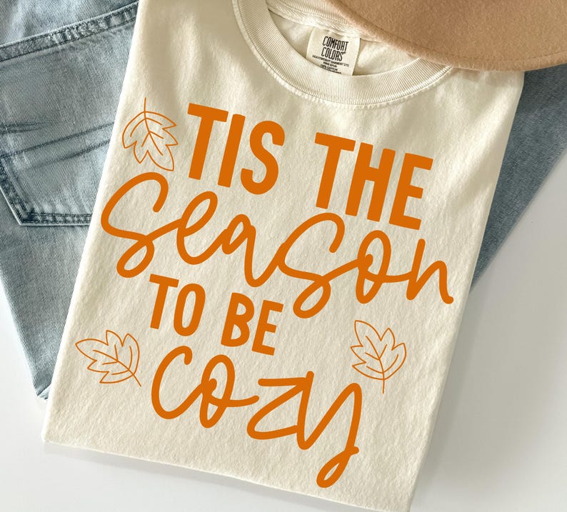 May include: A cream-coloured t-shirt featuring the orange text 'TIS THE SEASON TO BE COZY' printed on the front. The design includes simple orange leaf illustrations positioned near the text. The shirt has a crew neckline and a Comfort Colors label.