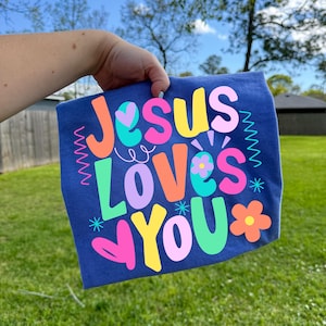 May include: A blue t-shirt with the words "Jesus Loves You" in colorful, playful lettering. The design includes flowers, stars, and decorative lines. The shirt is held up against a green grassy background.