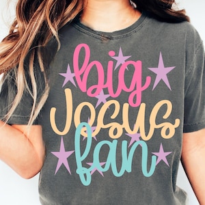 K&ouml;nnte beinhalten: Dunkelgraues T-Shirt mit dem Schriftzug "big Jesus fan" in Rosa, Pfirsich und T&uuml;rkis, akzentuiert mit lavendelfarbenen Sternen. Das Shirt ist ein l&auml;ssiger Stil.