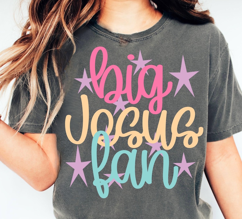 K&ouml;nnte beinhalten: Dunkelgraues T-Shirt mit dem Schriftzug "big Jesus fan" in Rosa, Pfirsich und T&uuml;rkis, akzentuiert mit lavendelfarbenen Sternen. Das Shirt ist ein l&auml;ssiger Stil.