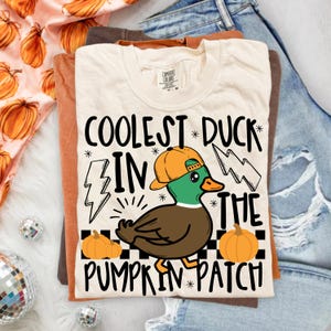 May include: A cream-colored t-shirt featuring a graphic of a duck wearing an orange baseball cap, surrounded by two lightning bolts and two orange pumpkins. The text reads COOLEST DUCK IN THE PUMPKIN PATCH, set against a black and white checkerboard.