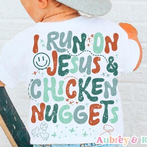 Puede incluir: Camiseta blanca con el texto "I RUN ON JESUS & CHICKEN NUGGETS" en varios colores. El diseño incluye una carita sonriente, estrellas e ilustraciones de pollitos. La marca "Aubrey & Rose" está en la parte inferior.
