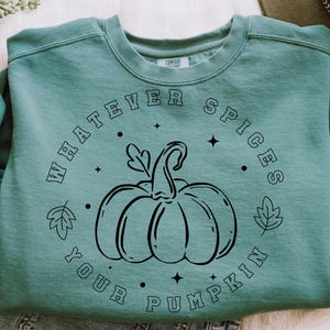 May include: A sage green crewneck sweatshirt featuring a black line art graphic of a pumpkin in the centre. The text "WHATEVER SPICES YOUR PUMPKIN" is printed in a circular arrangement around the pumpkin, accented by small leaf and star icons.