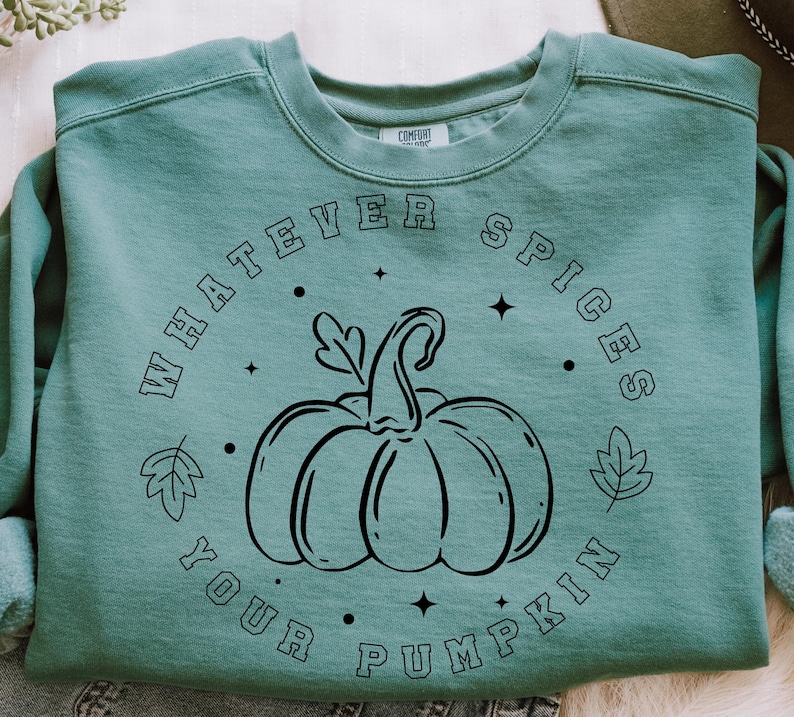 May include: A sage green crewneck sweatshirt featuring a black line art graphic of a pumpkin in the centre. The text "WHATEVER SPICES YOUR PUMPKIN" is printed in a circular arrangement around the pumpkin, accented by small leaf and star icons.