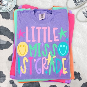 May include: A purple short-sleeve t-shirt with a crew neckline featuring the text Little Miss 1st Grade in pink, green, yellow, and light blue. The design includes yellow and blue smiley faces and various pink, green, and yellow star shapes.