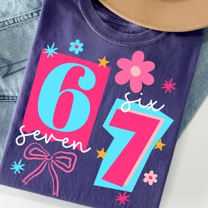 May include: A purple t-shirt with the numbers 6 and 7 in a pink and blue design, along with the words "six" and "seven" in white script. The shirt is decorated with pink flowers, stars, and a pink bow.