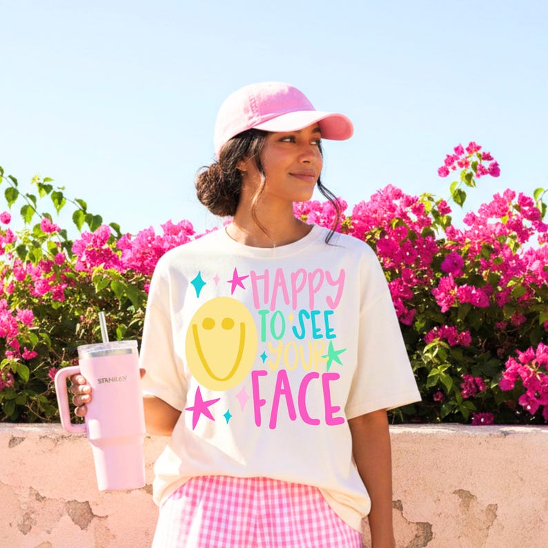 May include: A cream-colored t-shirt featuring a yellow smiley face and the text "HAPPY TO SEE YOUR FACE" in pink and teal, accented with star shapes. The shirt is paired with a pink baseball cap and a light pink insulated tumbler with a straw.