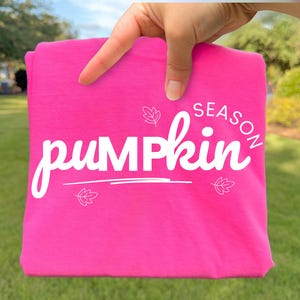 May include: A bright hot pink t-shirt featuring the white text "pumpkin SEASON" in a stylised script font. The design includes three small white line-art leaf illustrations positioned around the text, with a horizontal line underneath the word pumpkin.