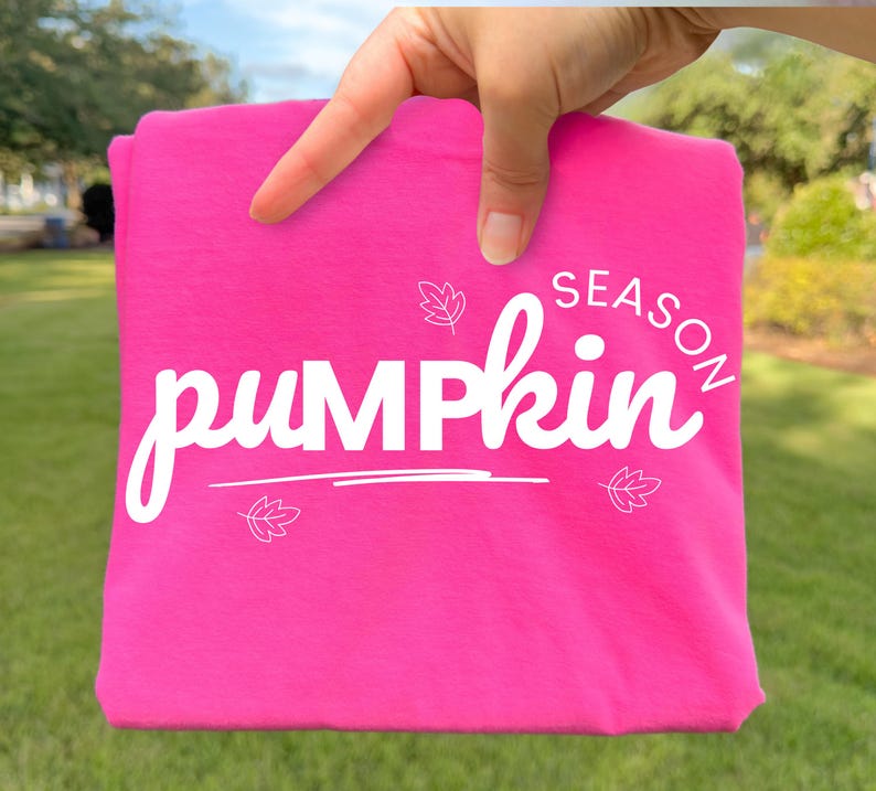 May include: A bright hot pink t-shirt featuring the white text "pumpkin SEASON" in a stylised script font. The design includes three small white line-art leaf illustrations positioned around the text, with a horizontal line underneath the word pumpkin.